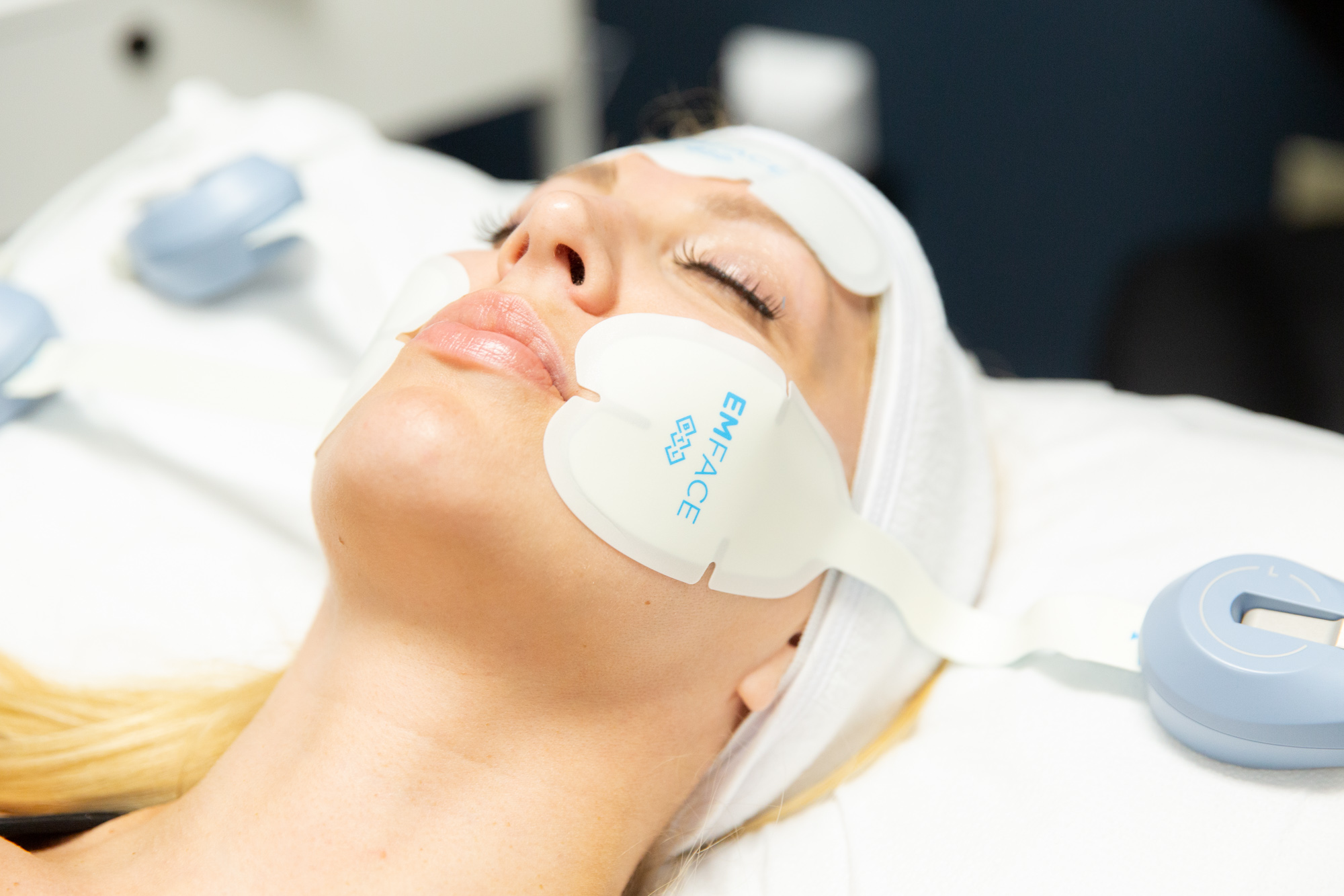 Female client relaxes with eyes closed on medical table during Emface treatment. Emface treatment patches are applied to her cheeks and forehead.