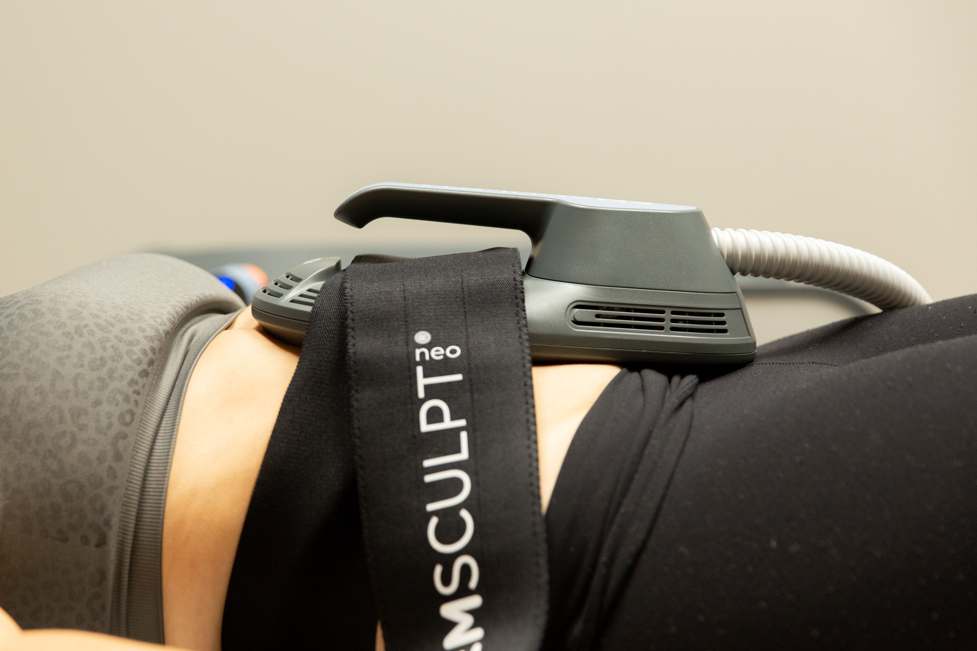 Close up image of woman receiving Emsculpt NEO treatment. The Emsculpt NEO applicator is pressed to the woman's stomach by the Emsculpt band, made of a soft, stretching material. The woman is lying down, image is shot from the side.