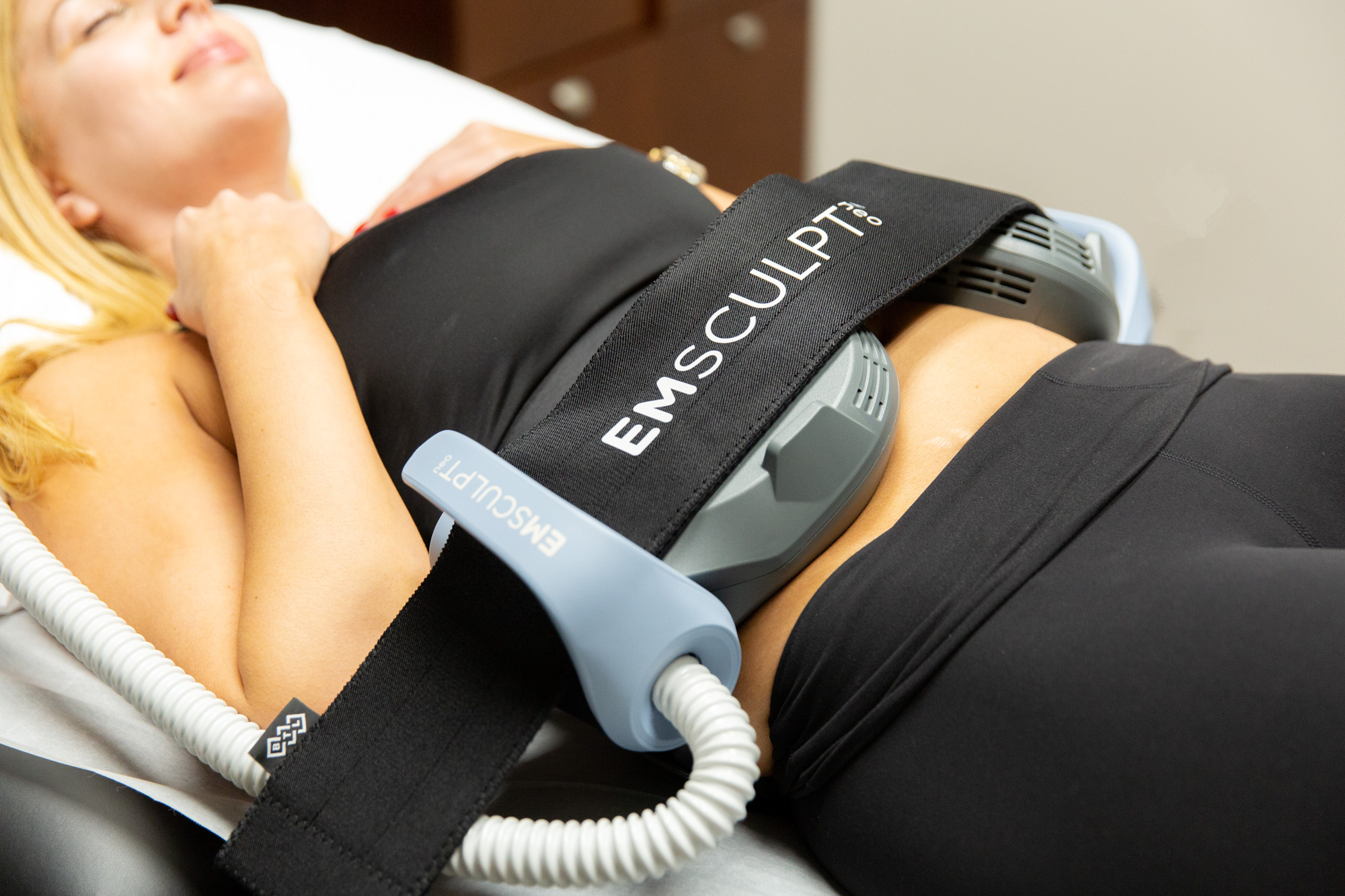 Two Emsculpt NEO applicators are attached to patient's stomach, one on right side and one on left. Patient is laying down with eyes closed during treatment.