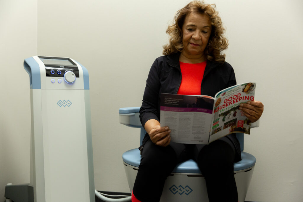 Female patient browses Good Housekeeping magazine while seated on Emsella chair, receiving treatment.