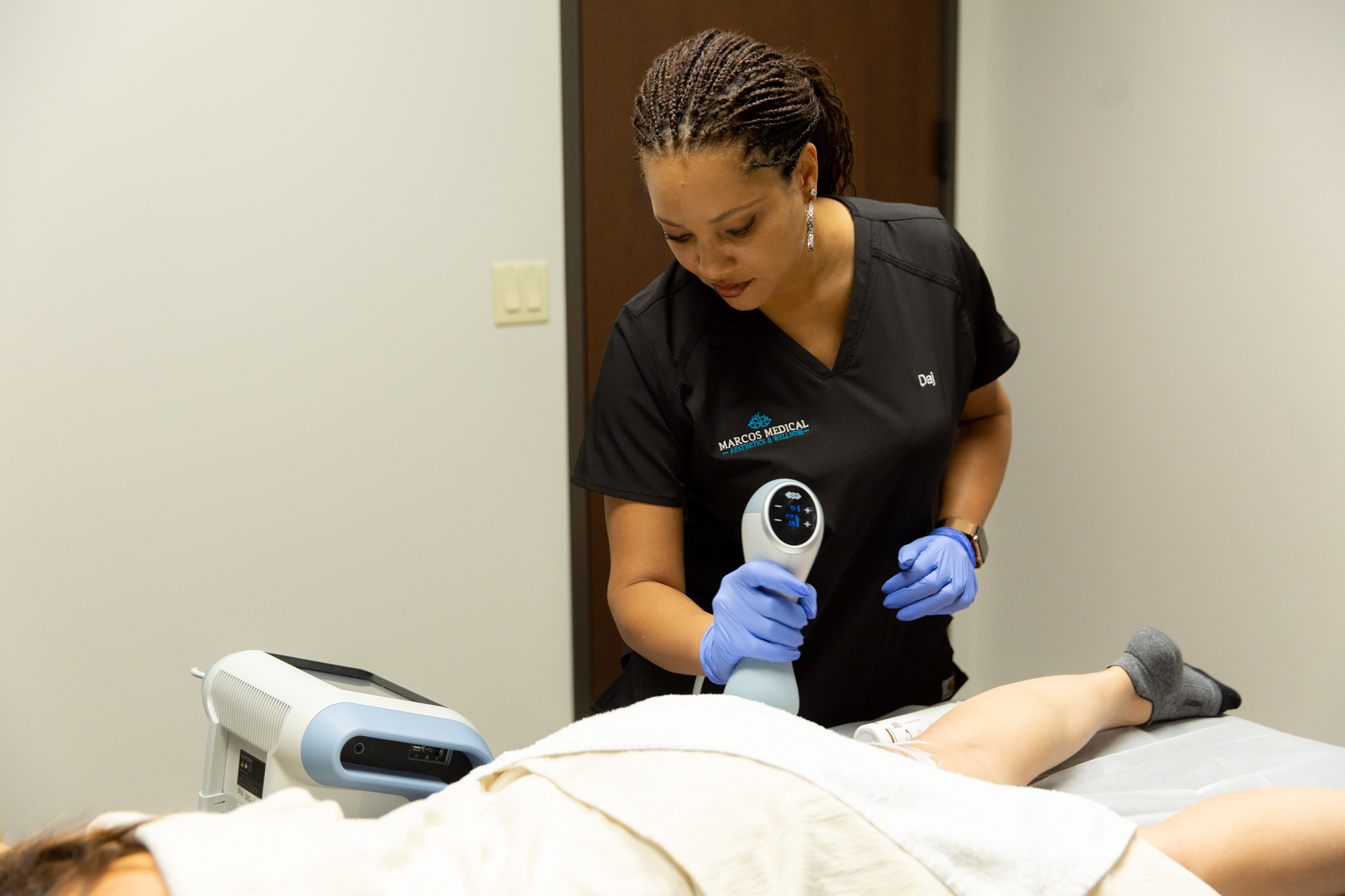 A female Marcos Medical Provider uses the Emtone handheld tool on a patient's thigh. The patient is laying face down on the medical table and is covered by a towel except for their legs. The Emtone machine is by the bedside of the patient.