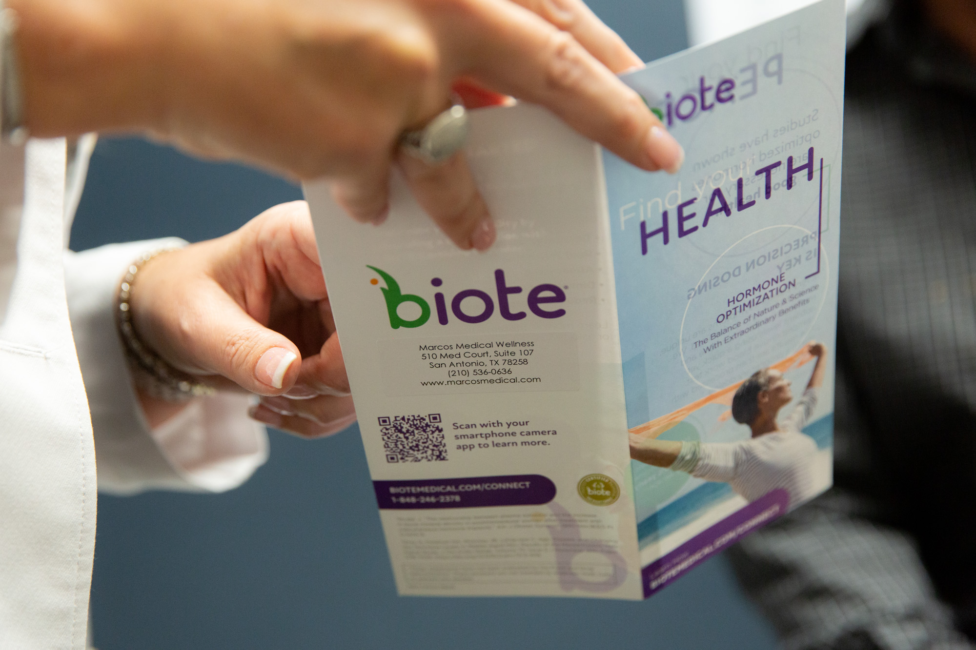 Marcos Medical Provider points to flyer describing hormone replacement therapy. The front of the flyer says Find your health - hormone optimization. The back of the flyer has the biote logo, Marcos Medical's address, and a QR code to scan with phone to learn more.