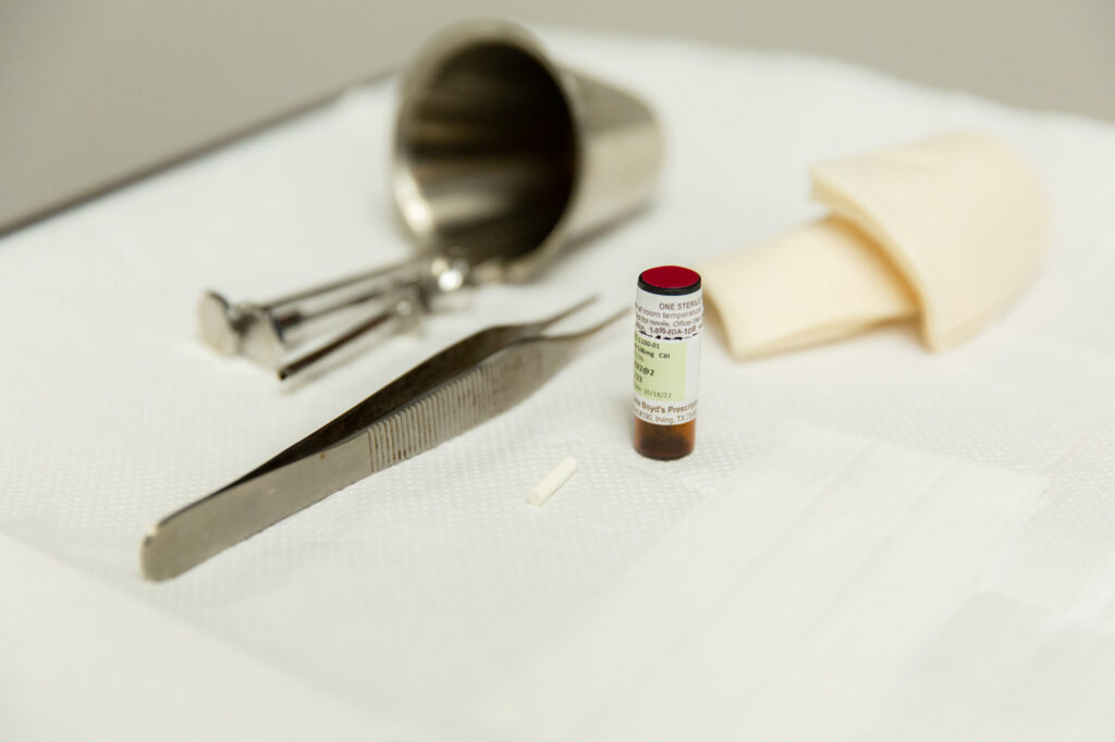 Tray table close up. Tweezers, square gauzes, hormone replacement pellet, small bottle, and metal cup holding two other metal medical instruments.