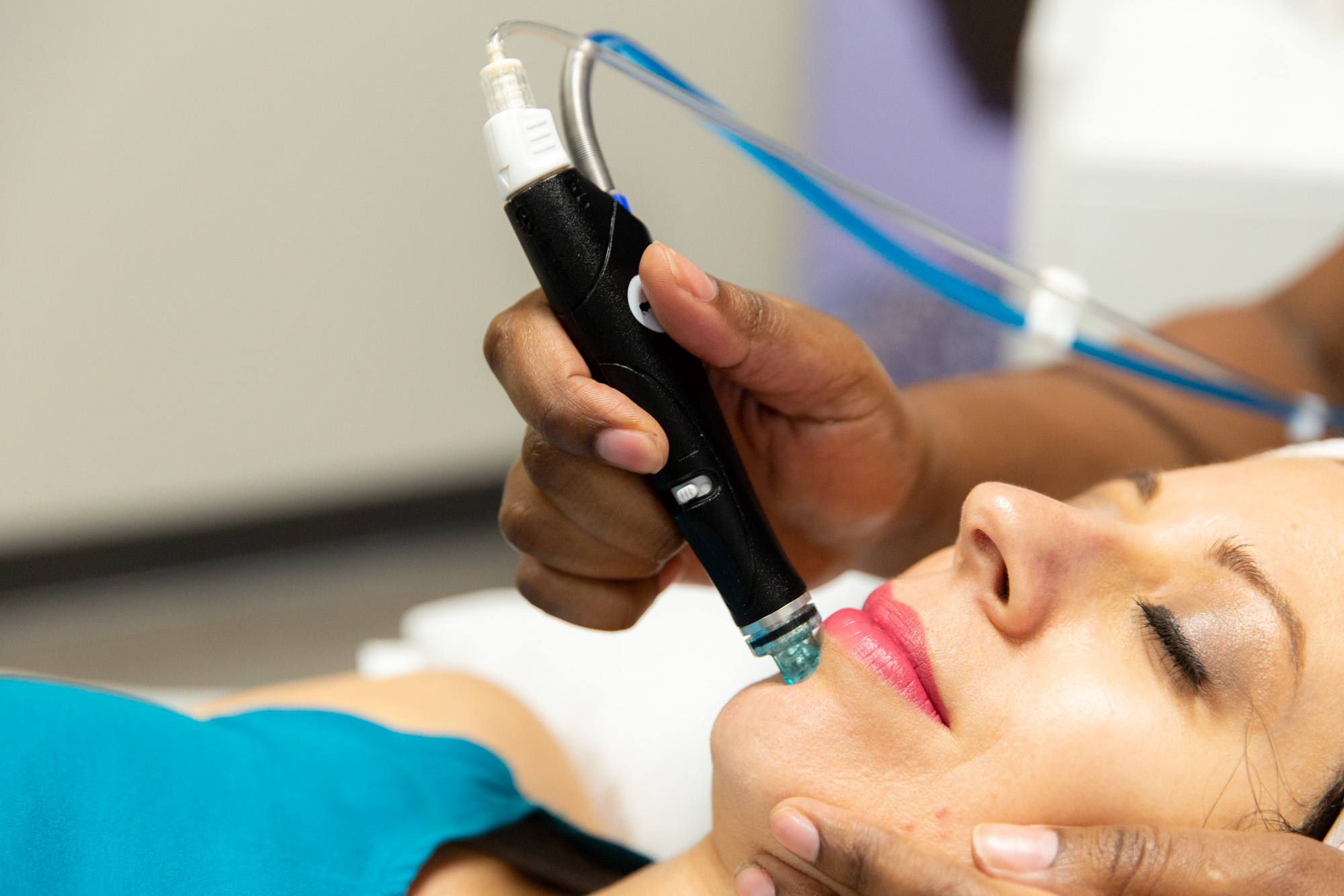 A Marcos Medical Provider applies the HydraFacial vacuum-based abrasion tool to a woman's chin during her facial. The provider is using her other hand to gently support the woman's face. The woman is laying down with her eyes closed.