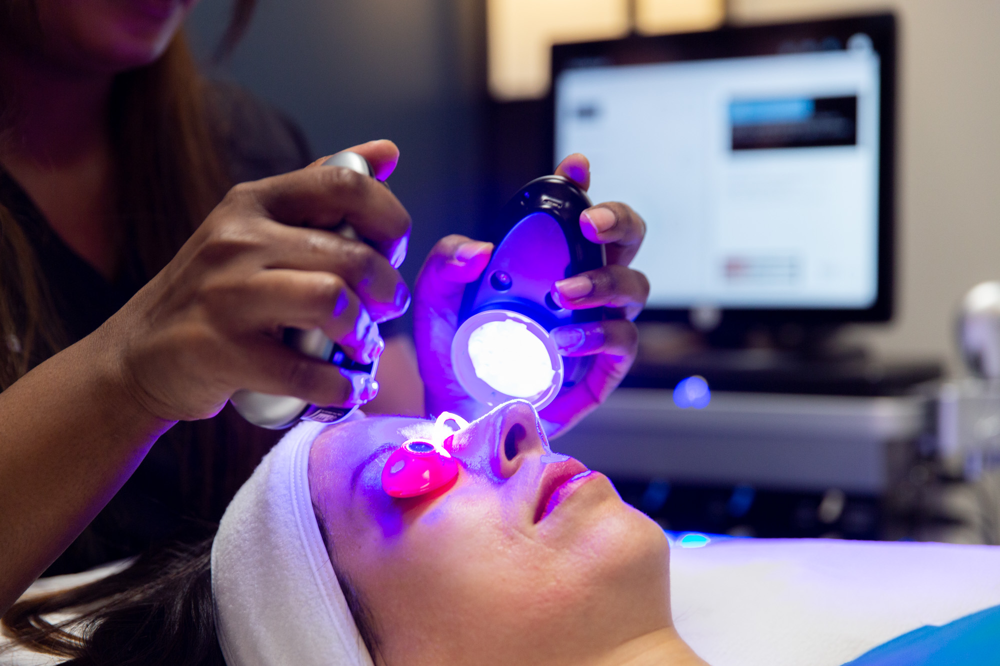 A Marcos Medical provider holds two handheld lights up to a female client's face during a HydraFacial. The client is wearing eye shields while laying down. A towel keeps her hair out of her face.