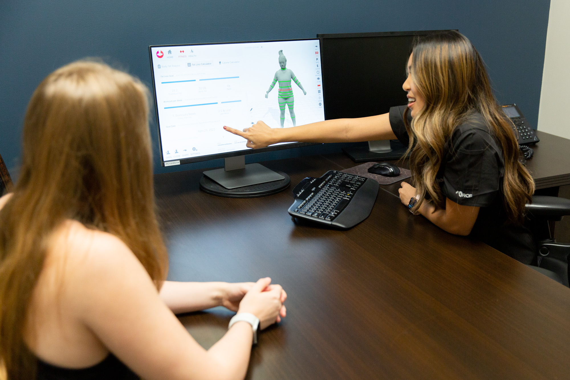 Marcos Medical provider points at a computer screen with a description of the Ideal Protein Plan. The screen also shows an image of a body. A female client follows along from the other side of the desk.