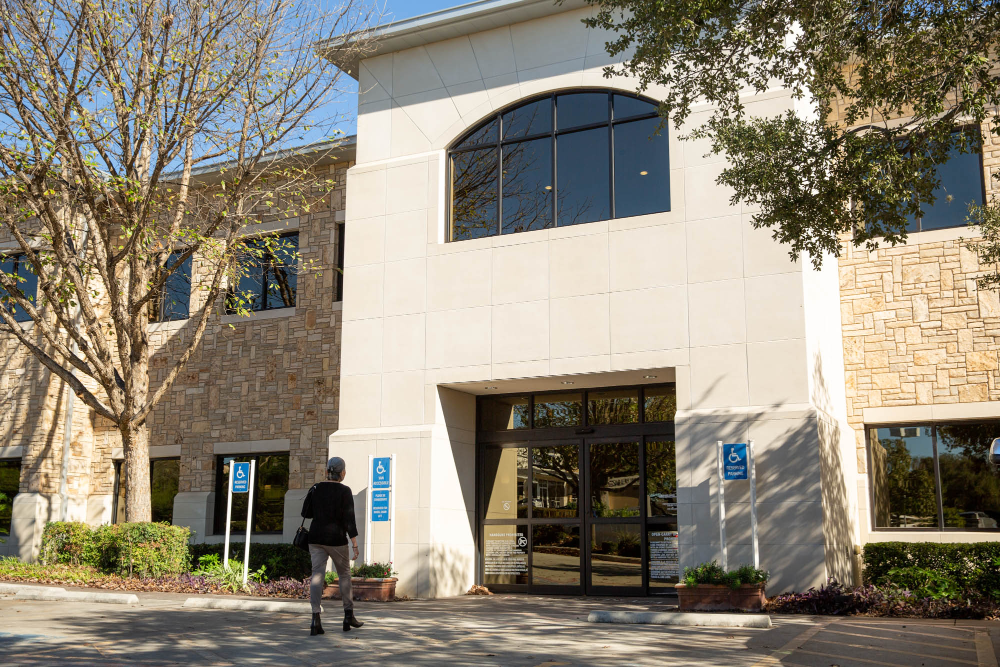 Exterior shot of Marcos Medical Care in San Antonio. The building is two stories made with light stone. Trees frame the entryway. A female client is walking to the doors.