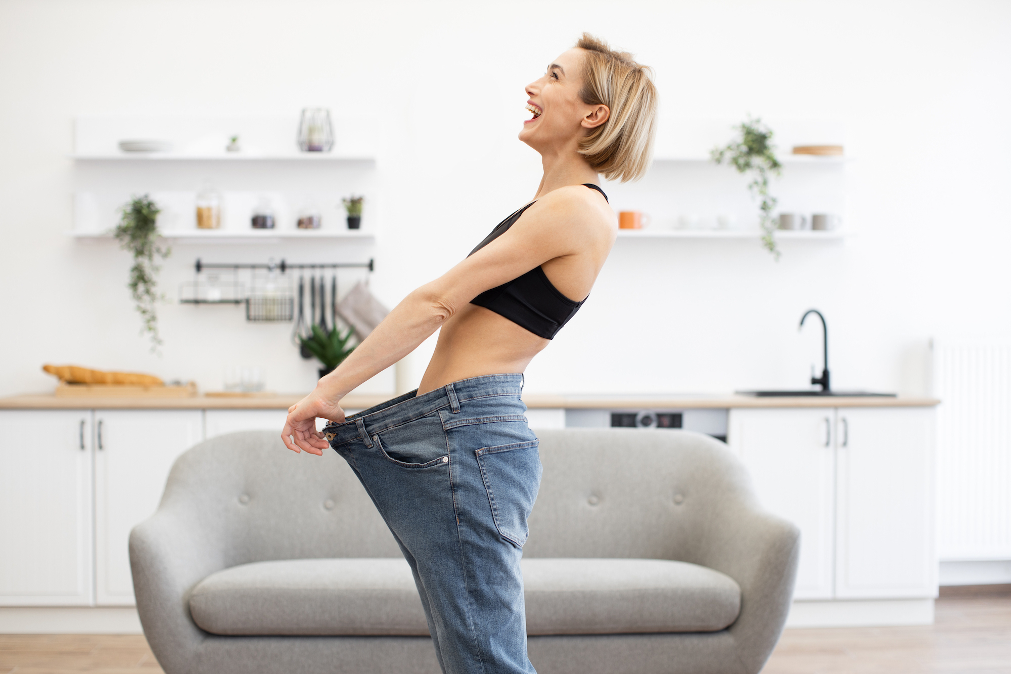 Happy woman in kitchen showing significant weight loss management in San Antonio while wearing oversized jeans. Success in dieting and fitness.