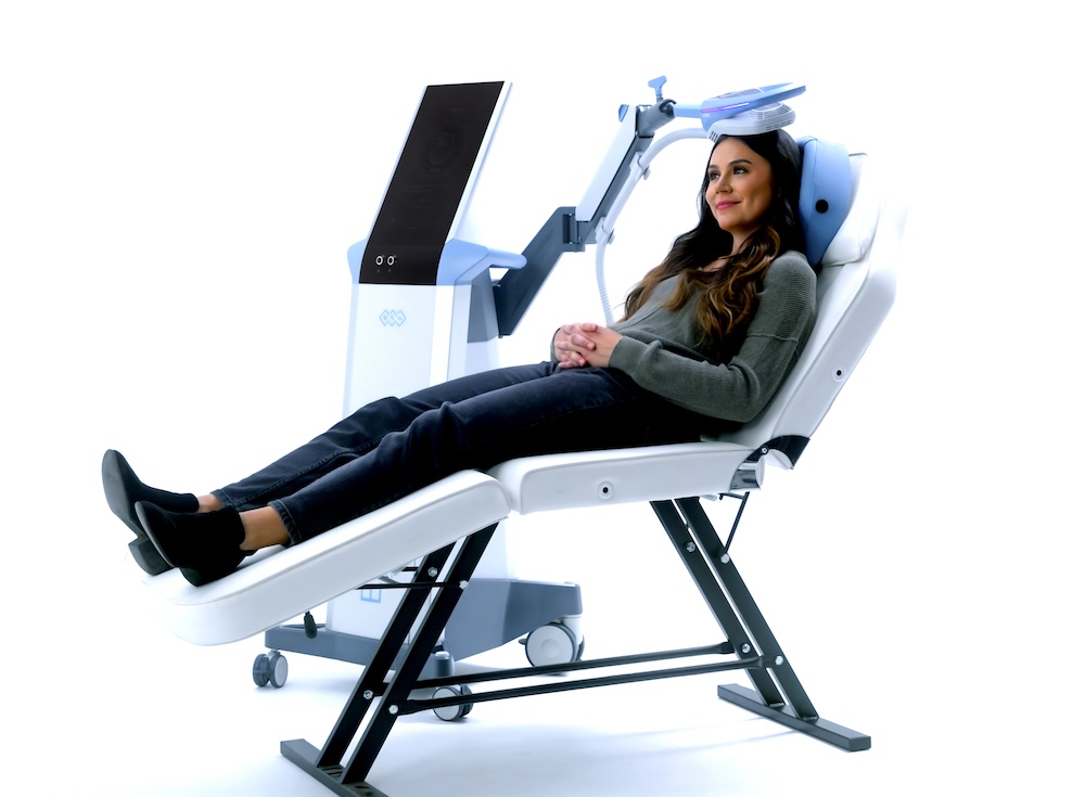 A woman with long, dark brown hair, a green long sleeve top, dark pants, and black boots, sitting on a treatment chair with the legs propped up and her hands folded over her lap, resting with the Exomind pad on her head at our wellness center in San Antonio.