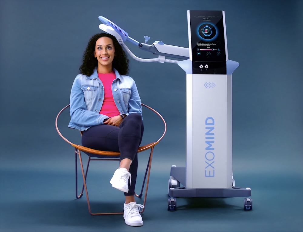 A young woman with curly, dark hair, wearing a jean jacket, pink shirt, dark blue skinny jeans, and white sneakers, sitting to the left of the Exomind machine, with the padded headpiece against the top of her head, receiving treatment at our wellness center in San Antonio.